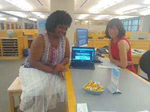 Two librarians seated at the Davis Family Library Research Desk, one using a stool