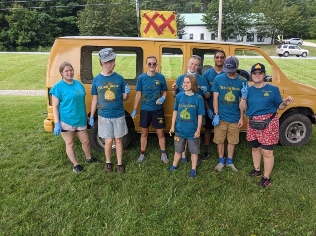 Eight individuals stand in front of a yellow van, smiling at the camera. They have yellow paint splattered on their blue Yellow House t shirts. 