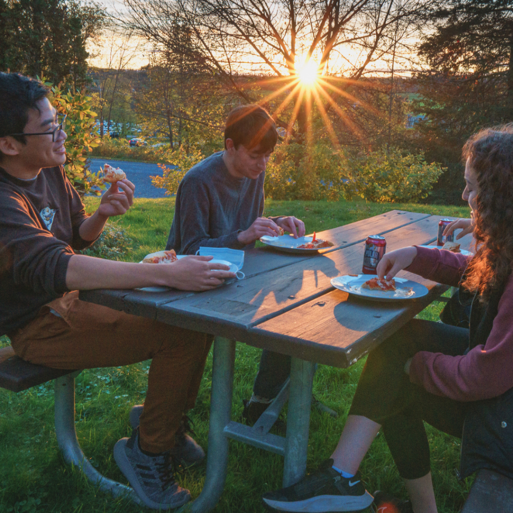Three students sitting outside the CCE enjoying pizza at sunset at the Privilege & Poverty pizza social.