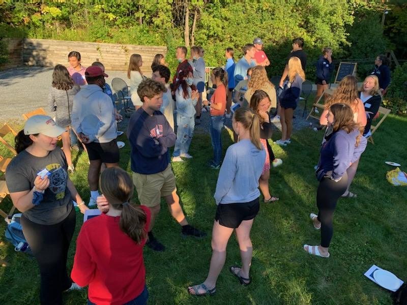 A group of Middlebury college students gather on the green grass outside the Center for Community Engagement. They're standing in pairs, talking with each other. 